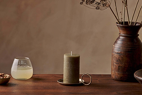 The Nkuku Rustic Soy Blend Pillar Candle in olive green sits on a brass holder atop a wooden table, flanked by a glass of pale yellow liquid and nuts on the left and a brown ceramic vase with dried flowers on the right.
