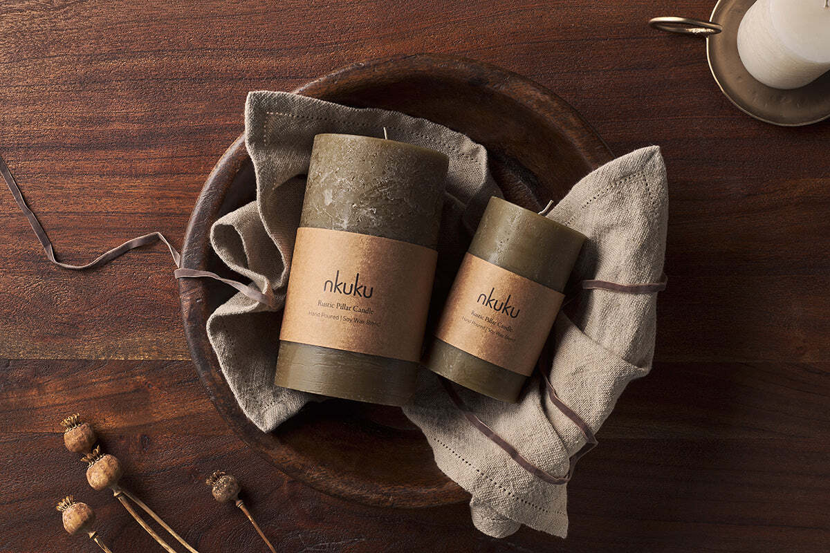 Two olive green Rustic Soy Blend Pillar Candles by Nkuku rest on a folded beige linen cloth inside a wooden bowl atop a dark wood table. Dried flower stems and a ribbon add to the warm, cozy scene.