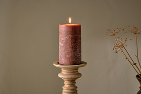 The nkuku Rustic Soy Blend Pillar Candle in Rust (Large) with a textured finish sits lit on a wooden holder against a beige wall, complemented by dried stems for a warm, minimalistic look.