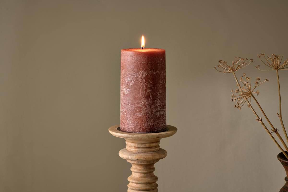 The nkuku Rustic Soy Blend Pillar Candle in Rust (Large) with a textured finish sits lit on a wooden holder against a beige wall, complemented by dried stems for a warm, minimalistic look.