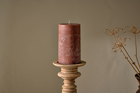 The nkuku Rustic Soy Blend Pillar Candle - Rust - Large, featuring a textured finish, is displayed on a wooden candlestick holder against a beige backdrop, with dried plant stems to the right for a natural accent.