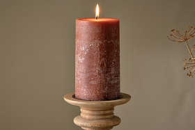 A lit nkuku Rustic Soy Blend Pillar Candle - Rust - Large with a textured finish sits on a round wooden holder against a plain beige background. A dry plant is faintly visible to the right.