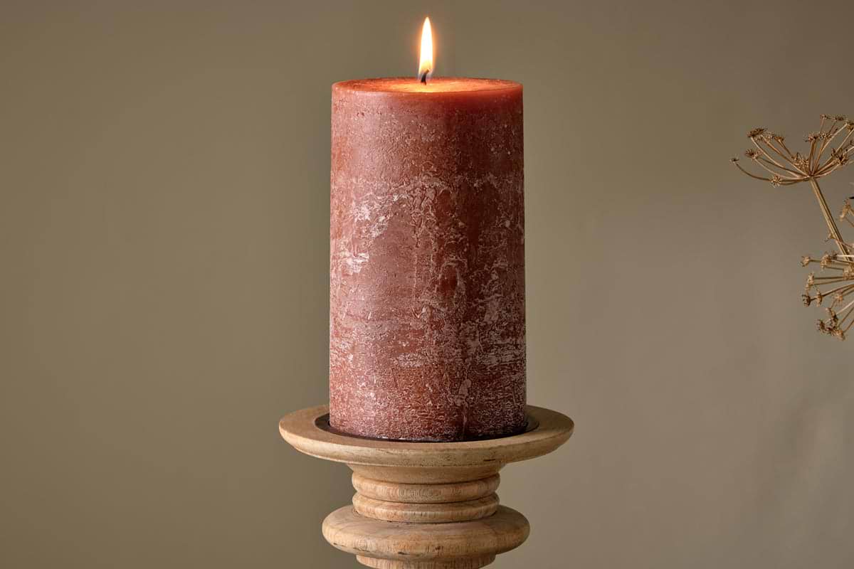 A lit nkuku Rustic Soy Blend Pillar Candle - Rust - Large with a textured finish sits on a round wooden holder against a plain beige background. A dry plant is faintly visible to the right.