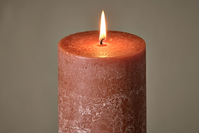 A close-up of the nkuku Rustic Soy Blend Pillar Candle in Rust (Large) showcases its textured reddish-brown wax, unique imperfections, and a warm flame glowing at the top against a softly blurred background.