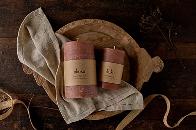Two nkuku Rustic Soy Blend Pillar Candles in Rust (Large) rest on a beige cloth atop a wooden tray. The setup, placed beside dried plant stems and tan ribbon, creates a warm, earthy ambiance on the dark wood surface.