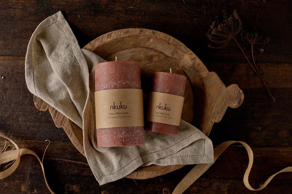Two nkuku Rustic Soy Blend Pillar Candles in Rust (Large) rest on a beige cloth atop a wooden tray. The setup, placed beside dried plant stems and tan ribbon, creates a warm, earthy ambiance on the dark wood surface.