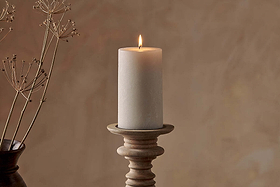 The Nkuku Rustic Soy Blend Pillar Candle in white (large) sits atop a light wood holder; a dark vase with dried stems is partly visible on the left, set against a softly textured beige background.