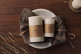 Two large white Rustic Soy Blend Pillar Candles by Nkuku with a textured finish rest on a gray stone plate atop a brown cloth on a wooden table. A third candle in a brass holder and dried grass complete the cozy, rustic scene.