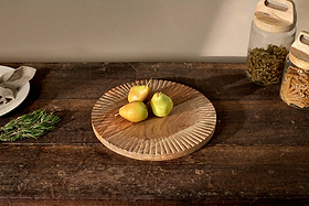 A nkuku Soria Chopping Board holds a hand-carved, round, ridged mango wood platter with four pears—three yellow, one green—at the center. To the right are glass jars of pasta; to the left, a plate with napkin, cutlery, and rosemary.