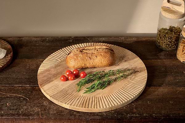 A round, hand-carved Soria Chopping Board by nkuku holds bread, cherry tomatoes, and fresh rosemary on a rustic table. Glass jars with dried herbs and pasta appear in the background as warm light casts gentle shadows.