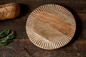 The nkuku Soria Chopping Board, hand carved with a textured rim, showcases natural wood grain and expert craftsmanship. It rests on a dark rustic surface beside fresh rosemary and a loaf of bread.