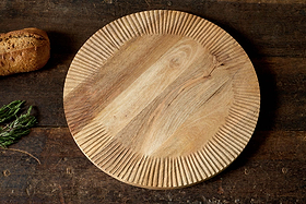The Soria Chopping Board by nkuku, hand carved from mango wood with radiating grooves and a natural grain pattern, sits on a rustic wooden table beside fresh rosemary and a loaf of bread.
