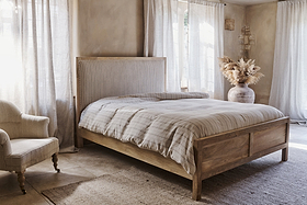 A cozy bedroom with nkuku’s Sanja Stripe Bed in natural solid mango wood, striped bedding, a cotton headboard, a cushioned armchair, sheer sunlit curtains, a vase of dried pampas grass, and neutral walls and floors.