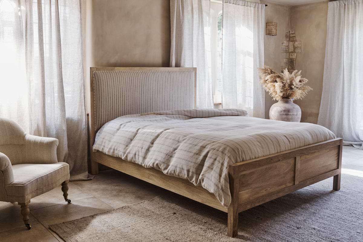 A cozy bedroom with nkuku’s Sanja Stripe Bed in natural solid mango wood, striped bedding, a cotton headboard, a cushioned armchair, sheer sunlit curtains, a vase of dried pampas grass, and neutral walls and floors.