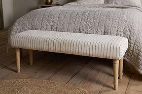 The nkuku Sanja Stripe Upholstered Bench in grey, with natural cotton upholstery and wooden legs, sits at the end of a bed with a textured light bedspread on wooden flooring next to a circular woven rug.