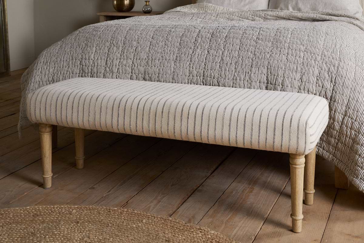 The nkuku Sanja Stripe Upholstered Bench in grey, with natural cotton upholstery and wooden legs, sits at the end of a bed with a textured light bedspread on wooden flooring next to a circular woven rug.