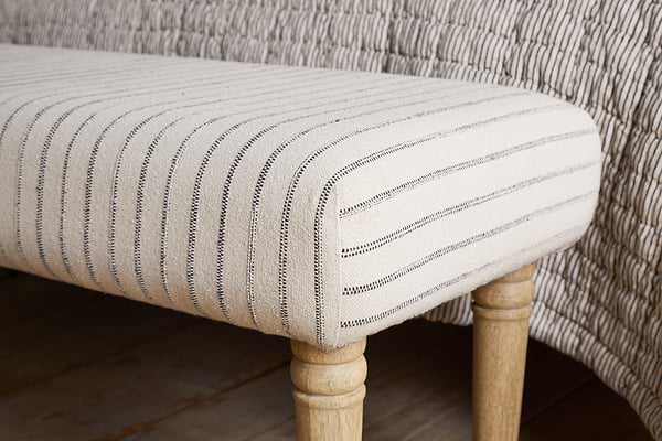 The Sanja Stripe Upholstered Bench - Grey by nkuku features natural cotton upholstery with black vertical stripes and light wooden legs, shown at the end of a bed beside a matching quilted blanket.