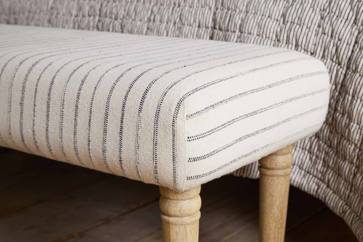 The Sanja Stripe Upholstered Bench - Grey by nkuku features natural cotton upholstery with black vertical stripes and light wooden legs, shown at the end of a bed beside a matching quilted blanket.