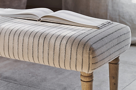 An open book rests on the nkuku Sanja Stripe Upholstered Bench in Grey, featuring natural cotton upholstery with slim dark stripes and wooden legs, placed at the end of a bed on a neutral-toned floor in a softly lit room.