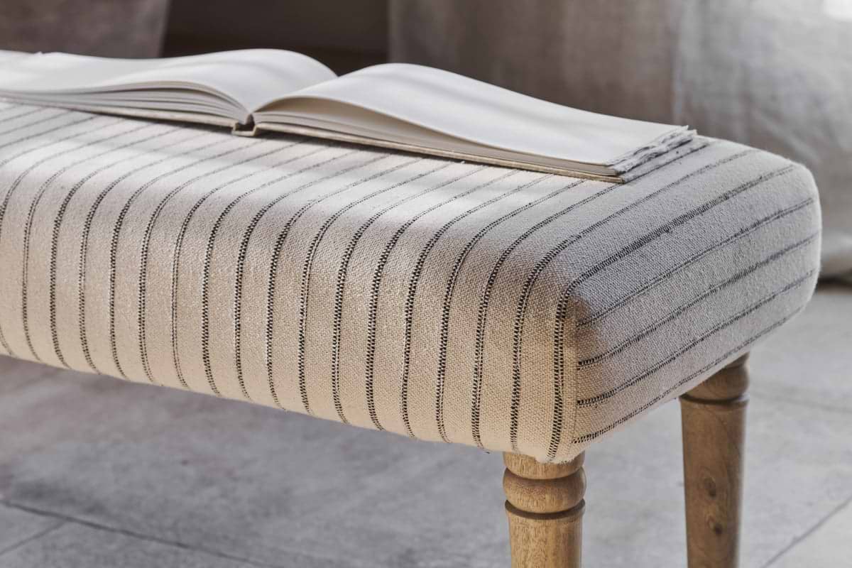 An open book rests on the nkuku Sanja Stripe Upholstered Bench in Grey, featuring natural cotton upholstery with slim dark stripes and wooden legs, placed at the end of a bed on a neutral-toned floor in a softly lit room.