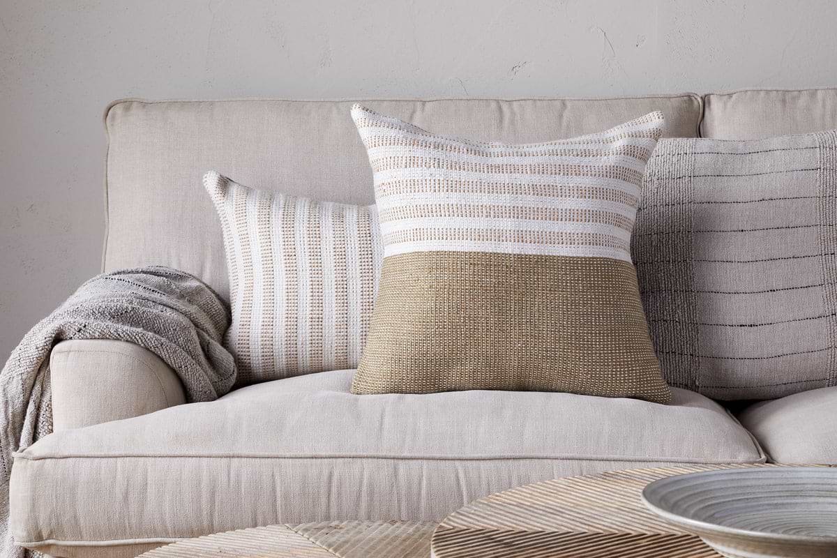 A beige sofa with a wool cushion cover and a folded gray blanket sits against a light wall. Two textured throw pillows, including the nkuku Samata Recycled Cushion Cover in Moss, add depth. A round woven coffee table is in the foreground.