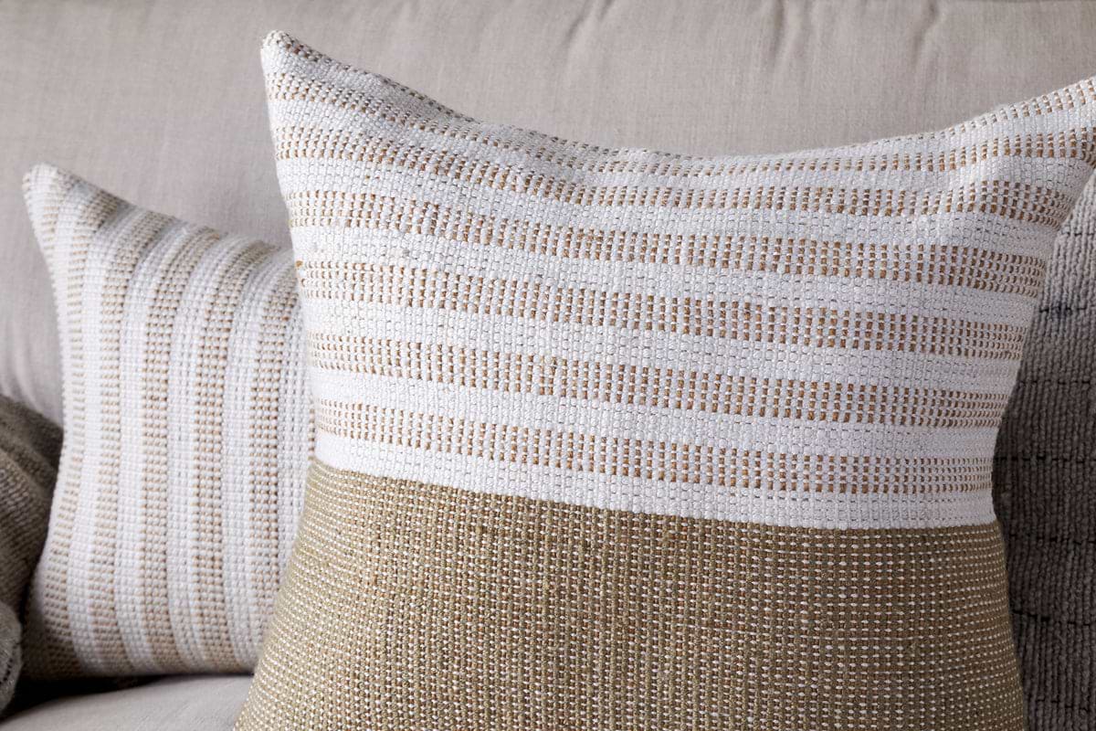 Two decorative throw pillows with woven textures, one in beige and white stripes and one with finer stripes, rest on a light sofa. Pair them with nkuku’s Samata Recycled Cushion Cover in Moss to enhance depth and style.