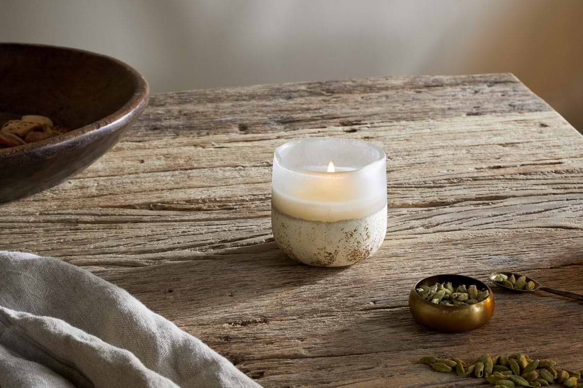 The nkuku Cardamom & Amber 1 Wick Scented Candle in a frosted glass holder rests on a rustic wooden table near linen, a brass bowl of cardamom pods, and the edge of a large wooden bowl.