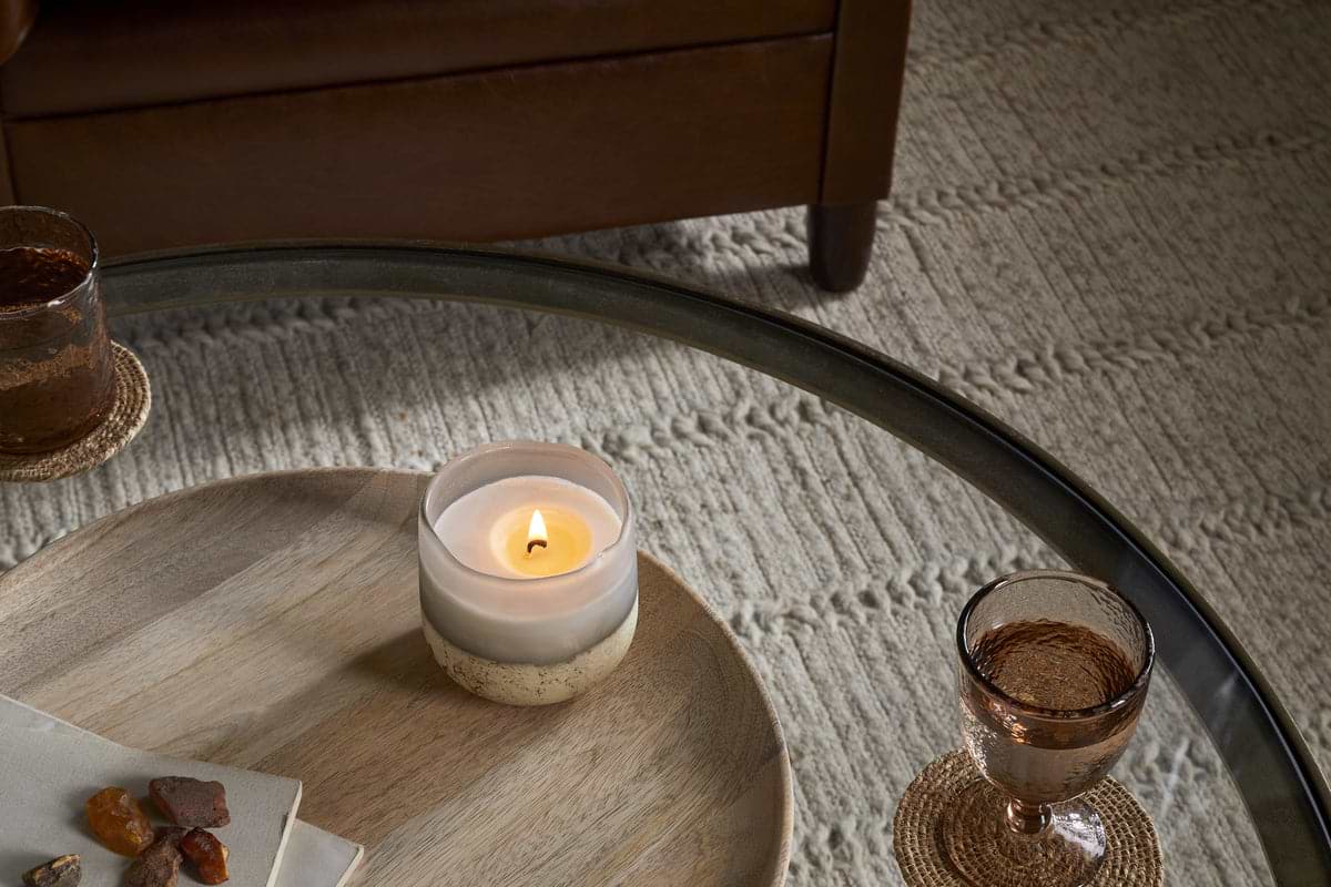 A Cardamom & Amber 1 Wick Scented Candle by nkuku in a two-toned holder sits on a round wooden tray with stones, next to two amber glasses on coasters, atop a glass coffee table over a textured beige rug.