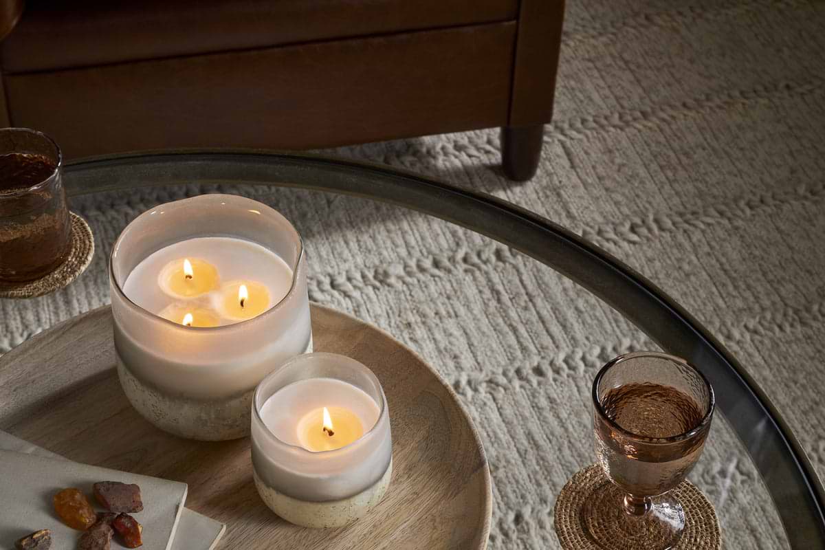 A glass coffee table displays two nkuku Cardamom & Amber 1 Wick Scented Candles in glass jars on a wooden tray, with a drink on a coaster and a folded napkin with stones, next to a beige textured rug and brown armchair.
