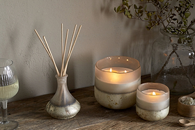 Two lit candles, the nkuku Sea Salt & Moss Reed Diffuser with sticks, a glass of white wine, and a jar of greenery are arranged on a rustic wooden table against a neutral wall.