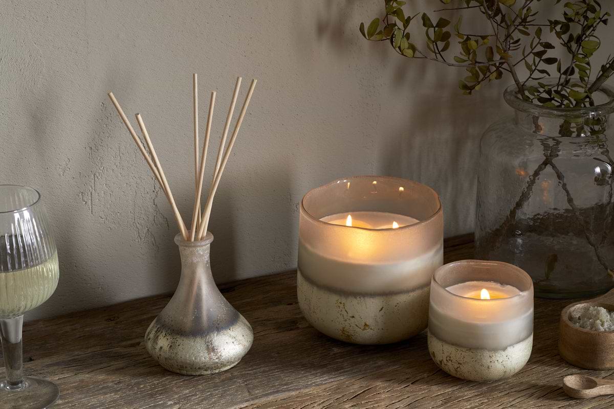 Two lit candles, the nkuku Sea Salt & Moss Reed Diffuser with sticks, a glass of white wine, and a jar of greenery are arranged on a rustic wooden table against a neutral wall.