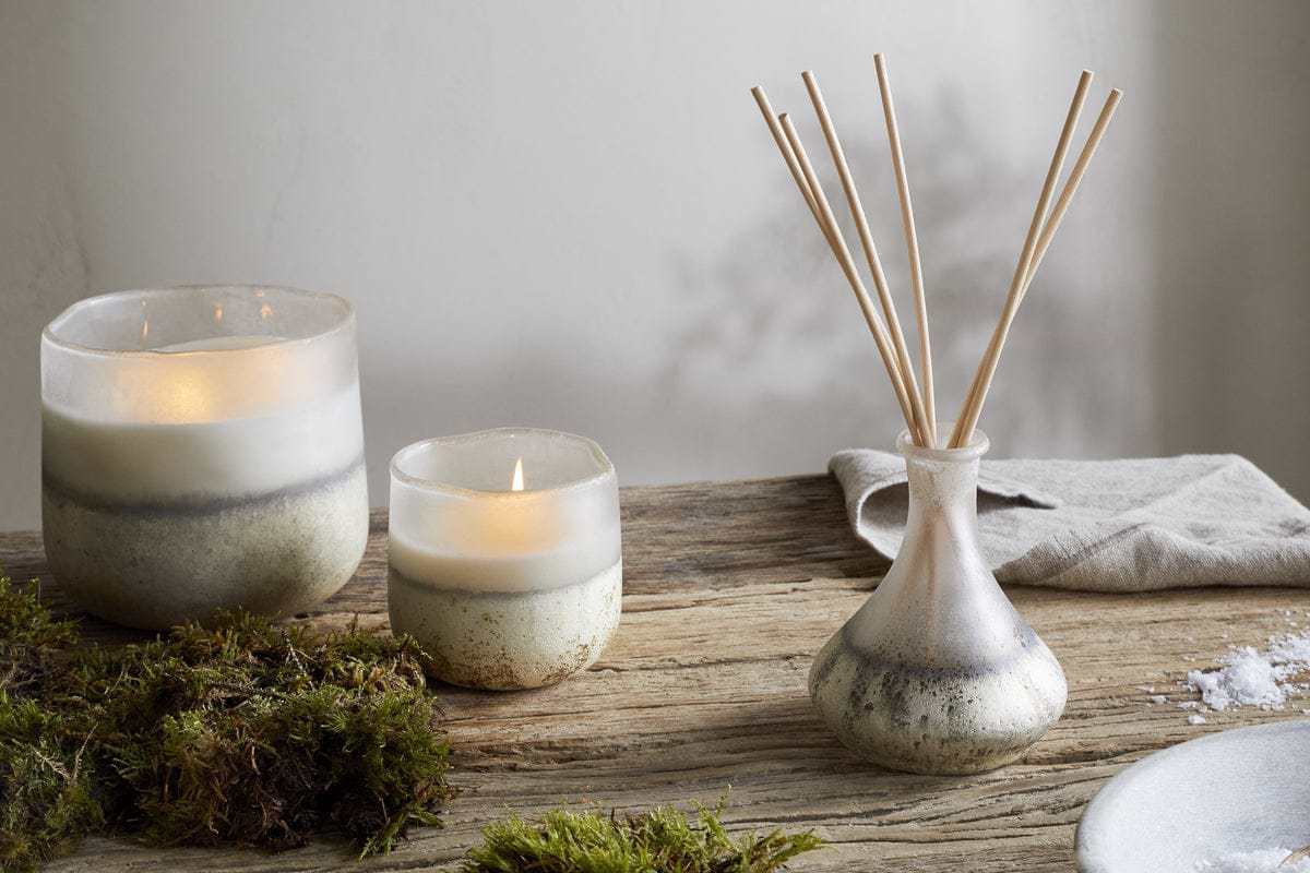 Two lit nkuku Sea Salt & Moss 1 Wick Scented Candles in ceramic holders rest on a rustic wooden table, accented with green moss and a folded beige cloth, creating a cozy ambiance filled with bespoke fragrance.