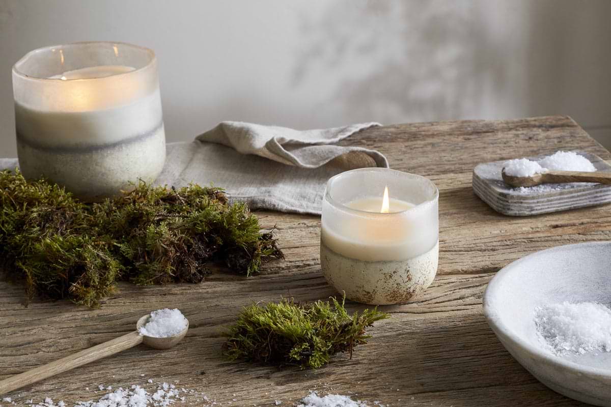 The nkuku Sea Salt & Moss 3 Wick Scented Candle in a glass holder fills a rustic wooden table with tranquil fragrance, surrounded by green moss, linen napkin, and wooden salt dishes—adding natural charm to any UK home.