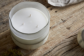 The nkuku Sea Salt & Moss 3 Wick Scented Candle, in a frosted glass jar, rests on a rustic wooden surface with a hint of light fabric in the background.