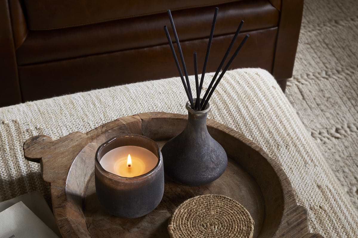 A lit candle, a small woven coaster, and the nkuku Cedarwood & Fig Reed Diffuser sit on a round wooden tray atop a textured beige ottoman, with a brown leather sofa in the background.