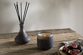 A lit candle in a dark glass holder, an nkuku Cedarwood & Fig Reed Diffuser with black reeds in a ceramic vase, and a wooden plate with sliced figs on a rustic wooden table.