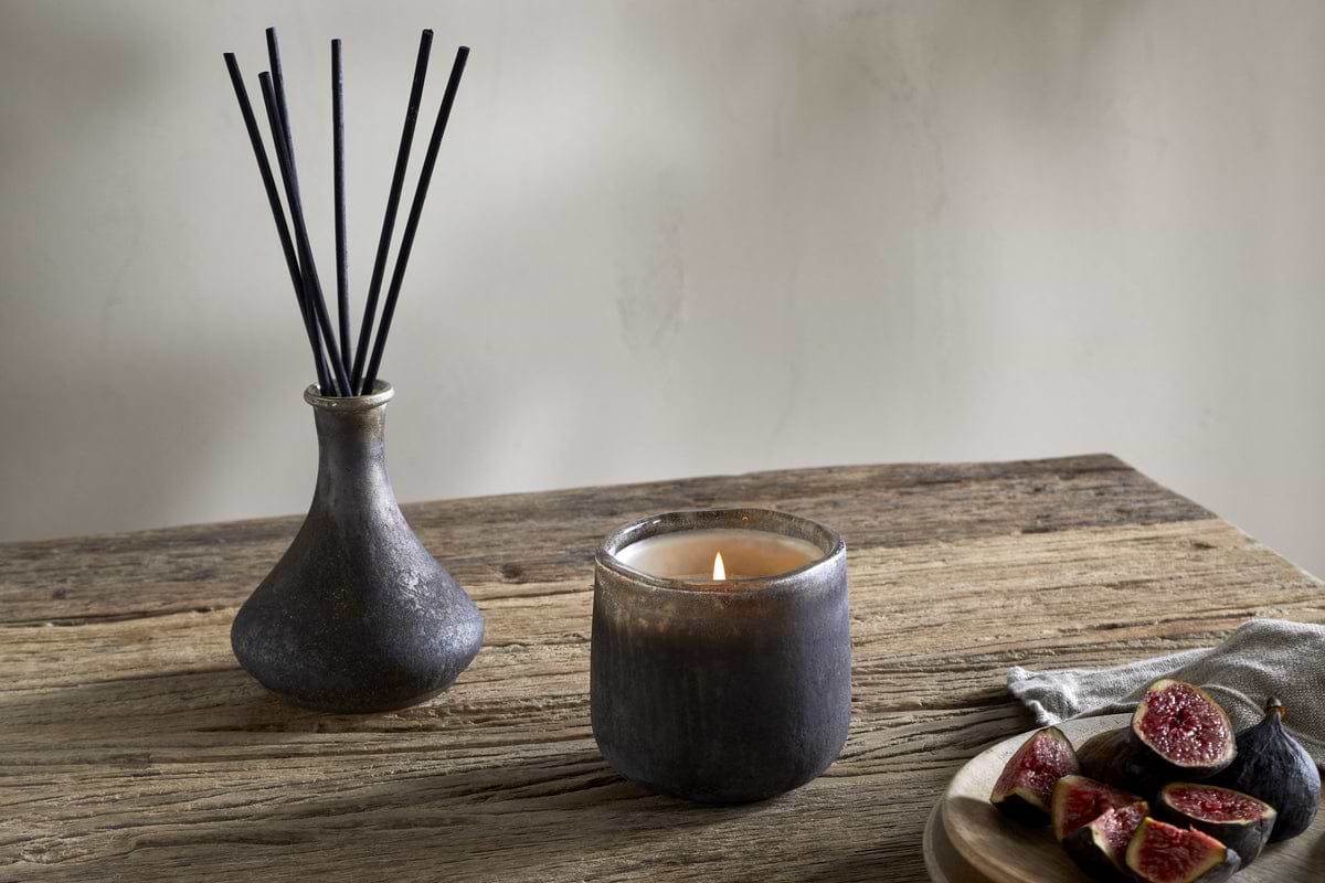 A lit candle in a dark glass holder, an nkuku Cedarwood & Fig Reed Diffuser with black reeds in a ceramic vase, and a wooden plate with sliced figs on a rustic wooden table.