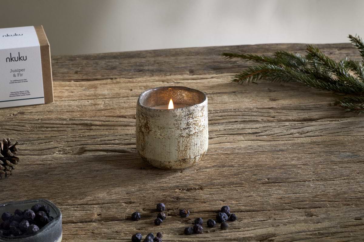 A Juniper & Fir 1 Wick Scented Candle by nkuku sits in a textured holder on a wooden table, surrounded by juniper berries, pinecone, evergreen sprig, and its labeled box—a bespoke fragrance in a reusable vessel.