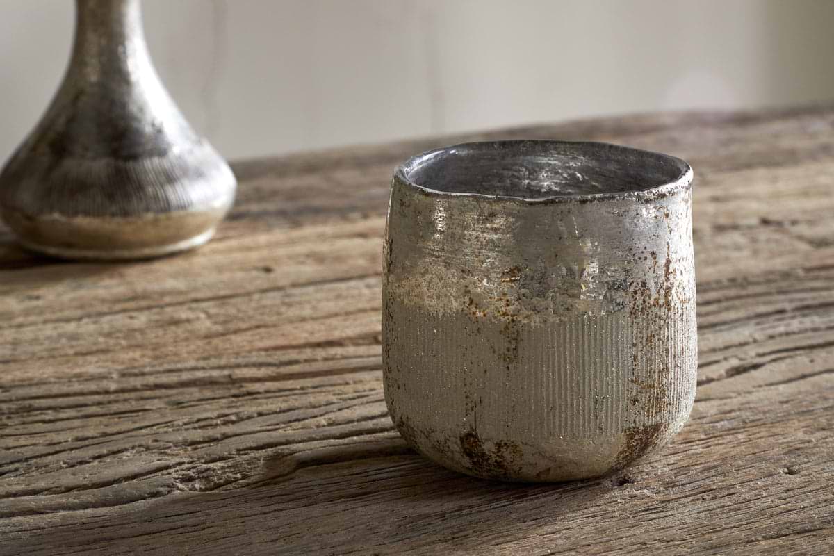 A Juniper & Fir 1 Wick Scented Candle by nkuku, in a weathered metallic holder with a textured finish, sits on a rustic wooden table. A blurred silver vase appears in the background.