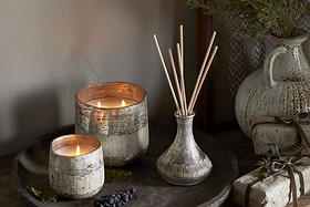 A rustic tray displays nkukus Juniper & Fir 3 Wick Scented Candle in a textured holder, a vase with reed diffuser sticks, juniper berries, greenery, and wrapped gifts on wood, with a ceramic jug behind.