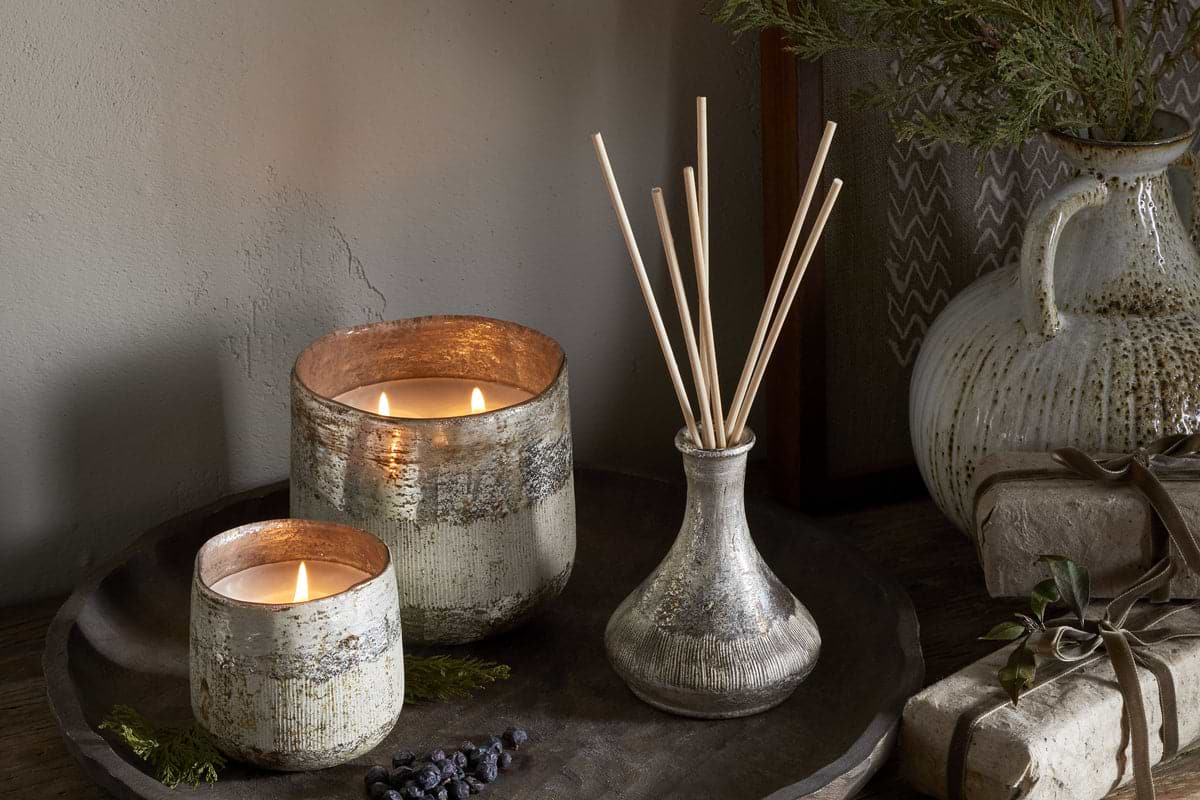 A rustic tray displays nkukus Juniper & Fir 3 Wick Scented Candle in a textured holder, a vase with reed diffuser sticks, juniper berries, greenery, and wrapped gifts on wood, with a ceramic jug behind.
