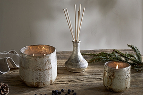 The nkuku Juniper & Fir 3 Wick Scented Candle, set on a rustic wooden table with pine branches, pine cone, and dried berries, fills the room with a cozy winter aroma.