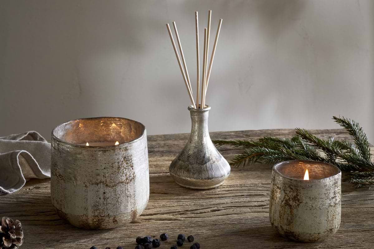 The nkuku Juniper & Fir 3 Wick Scented Candle, set on a rustic wooden table with pine branches, pine cone, and dried berries, fills the room with a cozy winter aroma.