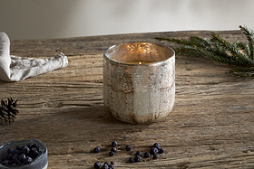 The nkuku Juniper & Fir 3 Wick Scented Candle glows in a rustic textured glass holder on a wooden table, surrounded by juniper berries, pine sprig, pine cone, and cloth—creating a cozy, natural winter atmosphere.
