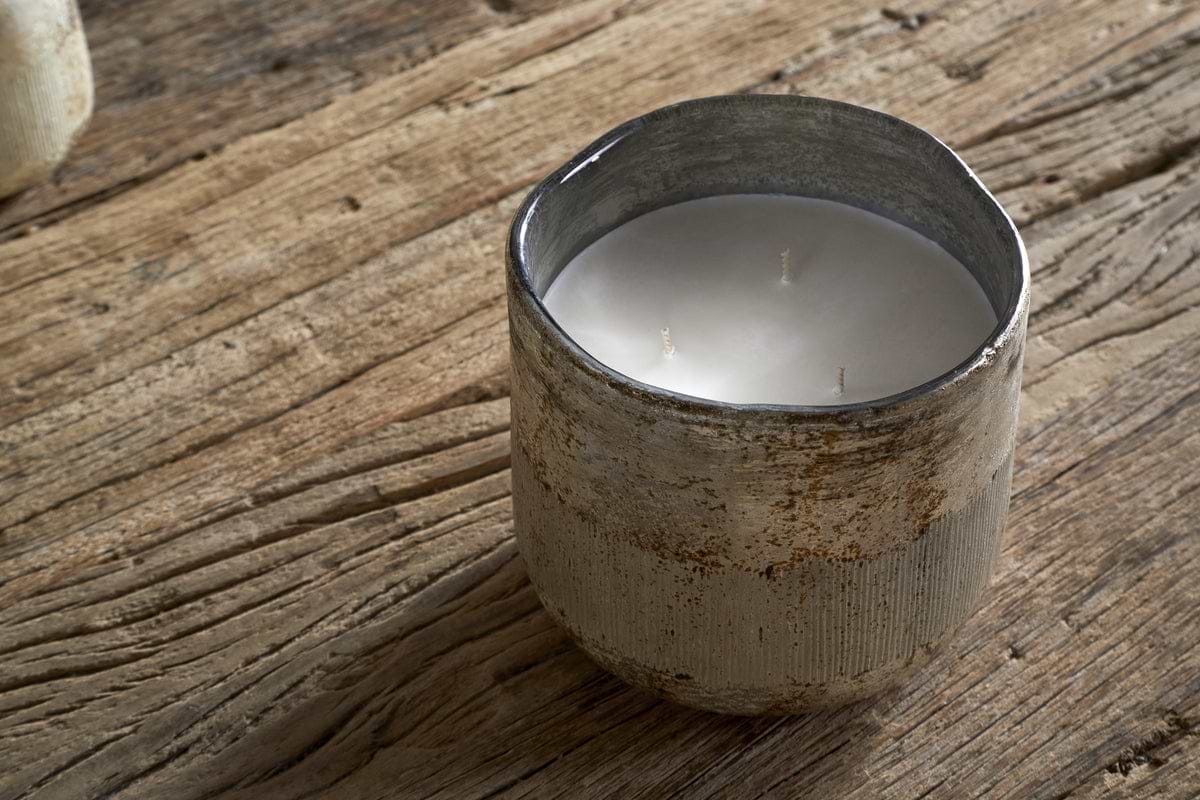 The nkuku Juniper & Fir 3 Wick Scented Candle, with a bespoke fragrance, sits in a rustic textured ceramic holder on a weathered wooden surface.
