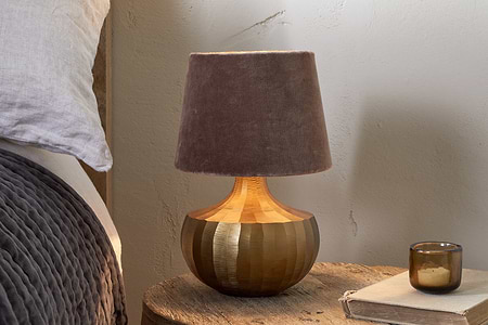 A bedside table displays the nkuku Sakiti Cast Aluminium Table Lamp in antique brass with a soft brown velvet shade, beside a small amber glass candle holder and old book. The gray quilted edge of a bed is visible on the left.