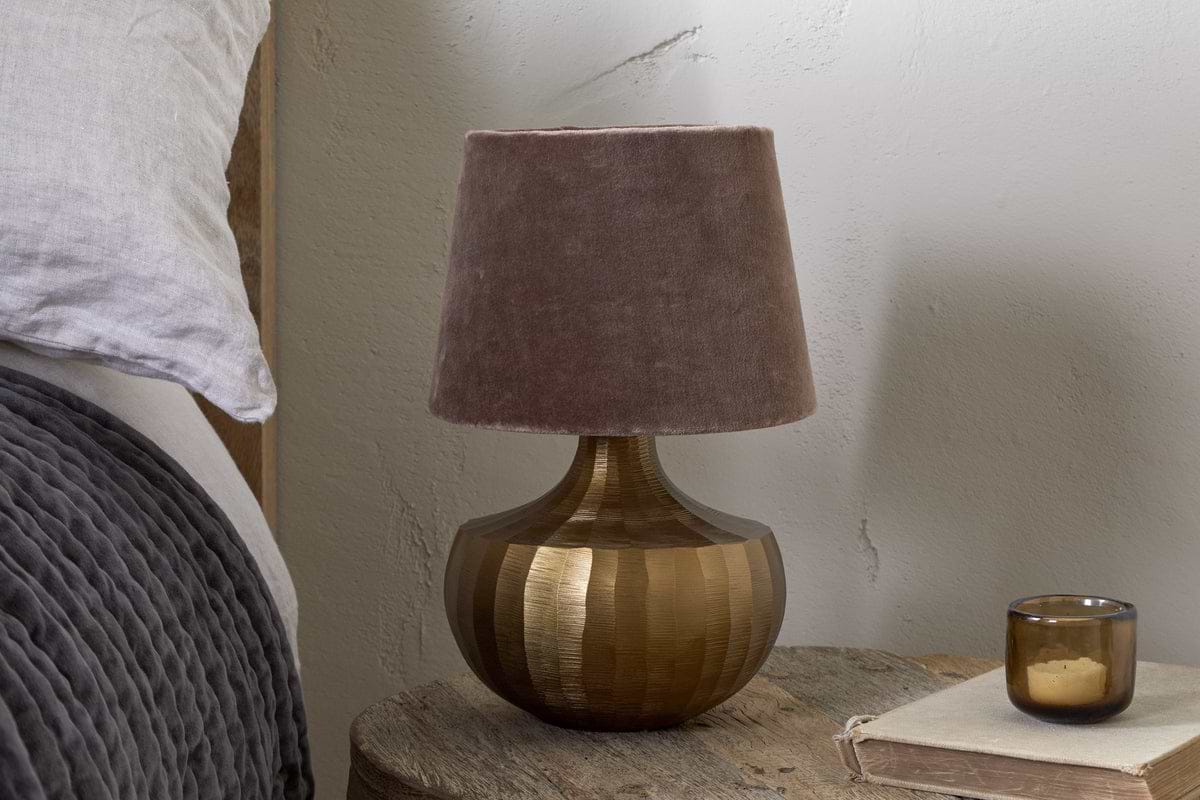 The Sakiti Cast Aluminium Table Lamp in Antique Brass by nkuku, featuring a textured round base and soft brown velvet shade, sits on a rustic nightstand beside gray bedding, with a closed book and brown glass candle holder nearby.