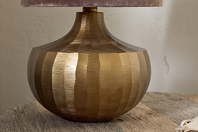 The nkuku Sakiti Cast Aluminium Table Lamp in Antique Brass features a sculptural, ribbed round base with vertical textured stripes, shown here on a rustic wooden surface with a partial view of its soft beige lampshade.