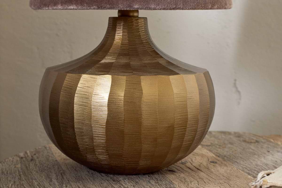 The nkuku Sakiti Cast Aluminium Table Lamp in Antique Brass features a sculptural, ribbed round base with vertical textured stripes, shown here on a rustic wooden surface with a partial view of its soft beige lampshade.
