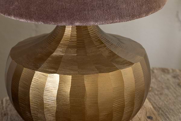 Close-up of the Sakiti Cast Aluminium Table Lamp by nkuku in antique brass, featuring a textured base with vertical ridges and topped with a soft, plush mauve lampshade on a rustic wooden surface.